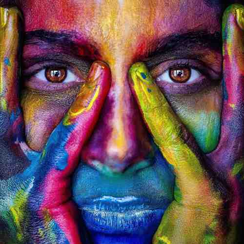 headshot of a woman holding her hands over her face and covered in colourful paint