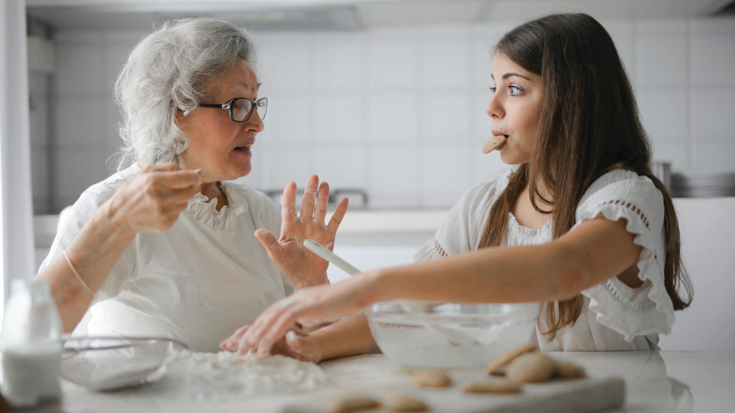 Calm senior woman and teenage girl in casual clothes looking at each other and talking while eating cookies and cooking pastry in contemporary kitchen at home