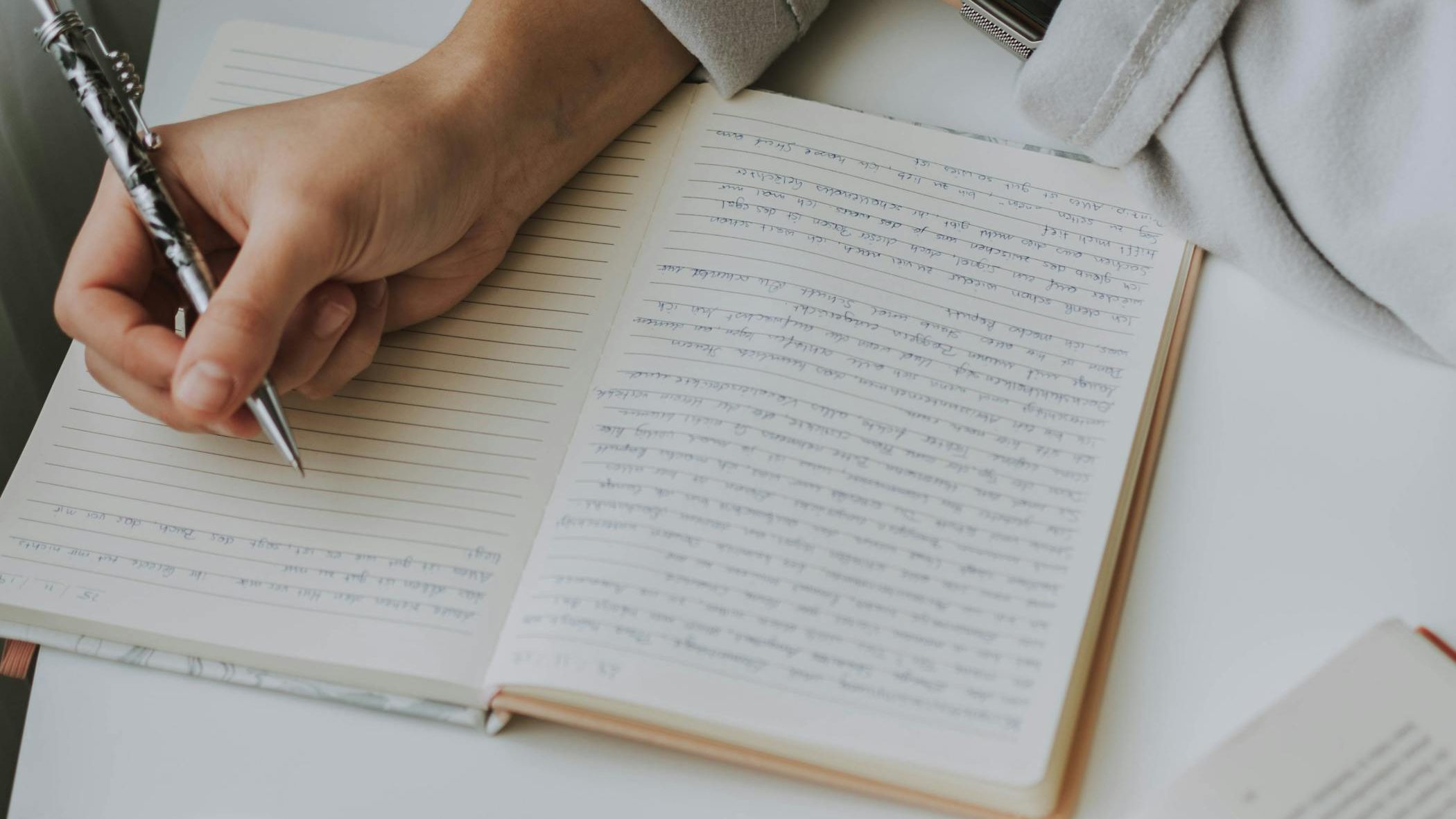 A person writing in a notebook with a cup of coffee and an open book on a white table. Handwriting Assessments and Occupational Therapy in Cornwall & Devon