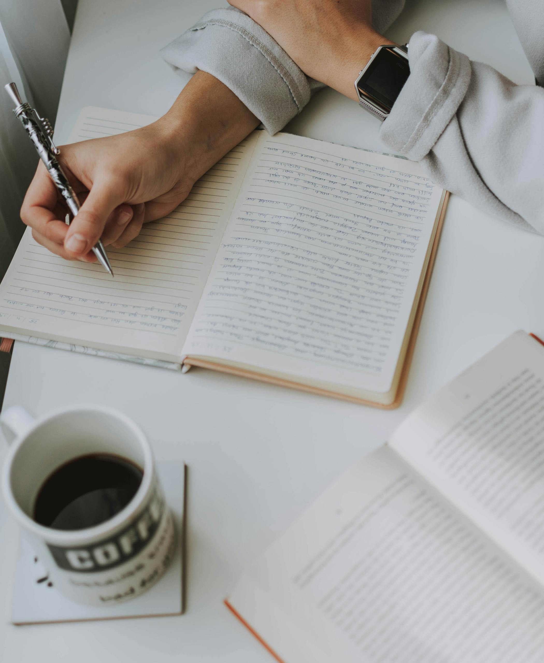 A person writing in a notebook with a cup of coffee and an open book on a white table. Handwriting Assessments and Occupational Therapy in Cornwall & Devon